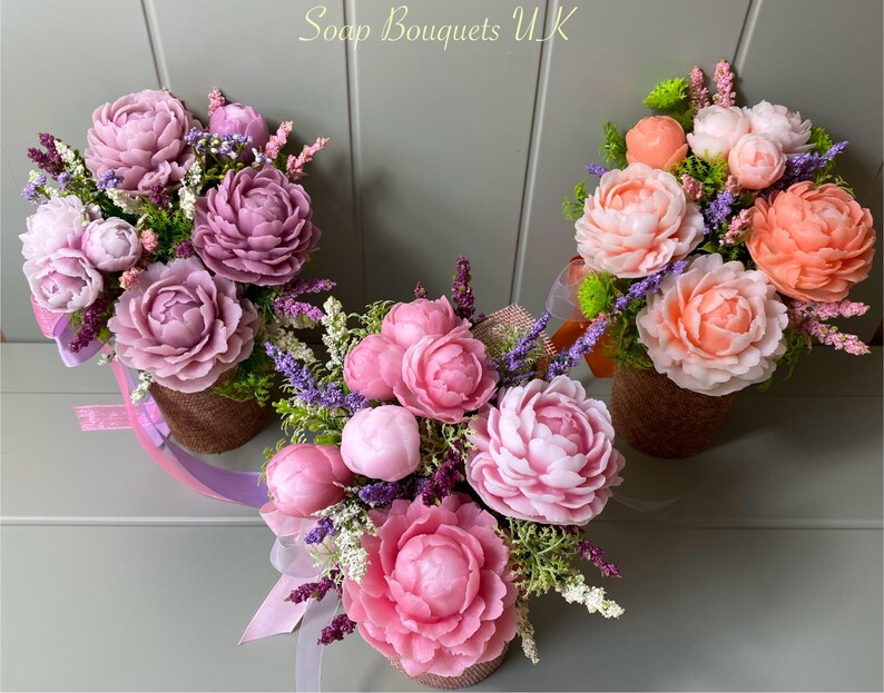 May include: Three soap flower bouquets in different shades of pink and peach. Each bouquet is arranged in a small brown basket and features a variety of flowers, including peonies, lavender, and greenery. The bouquets are arranged on a gray background.