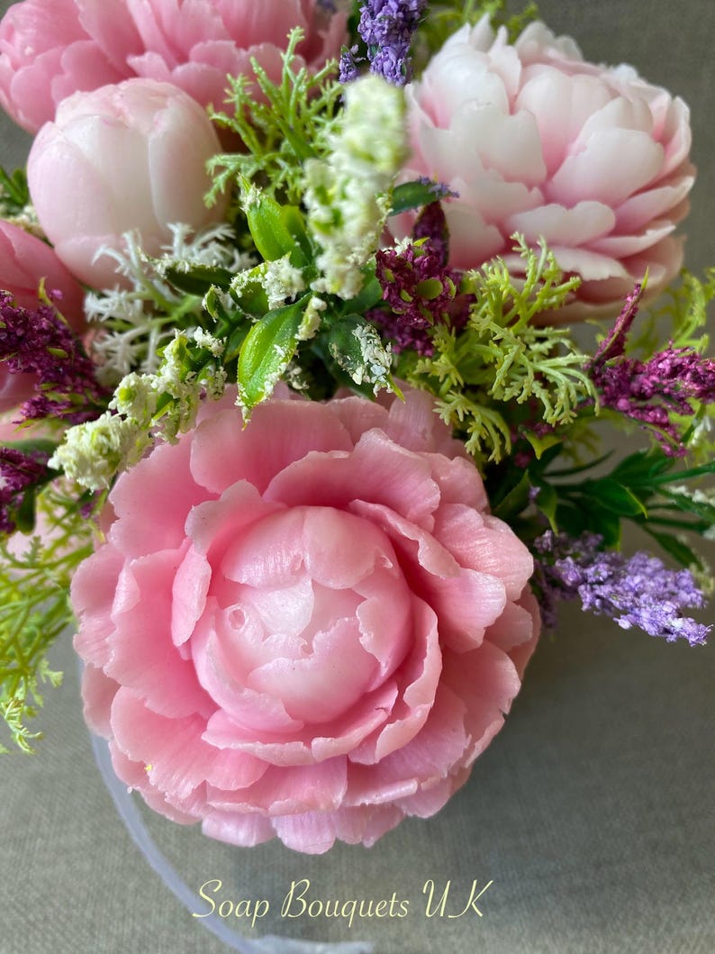 May include: A bouquet of pink soap flowers with green leaves and purple accents. The flowers are arranged in a clear vase. The text "Soap Bouquets UK" is visible at the bottom of the image.