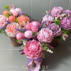May include: Three bouquets of pink and peach soap flowers arranged in brown pots. The flowers are made to look like peonies and are surrounded by green leaves and purple lavender.