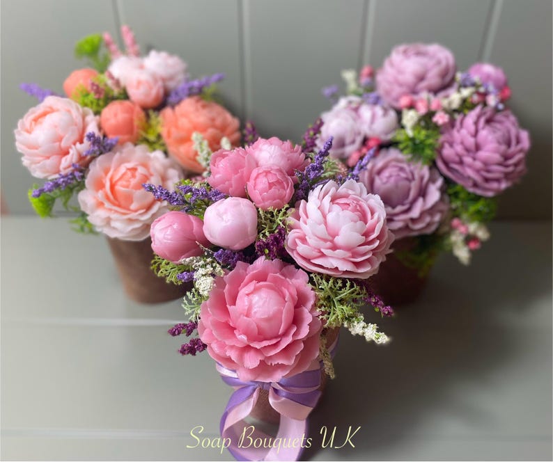 May include: Three bouquets of pink and peach soap flowers arranged in brown pots. The flowers are made to look like peonies and are surrounded by green leaves and purple lavender.