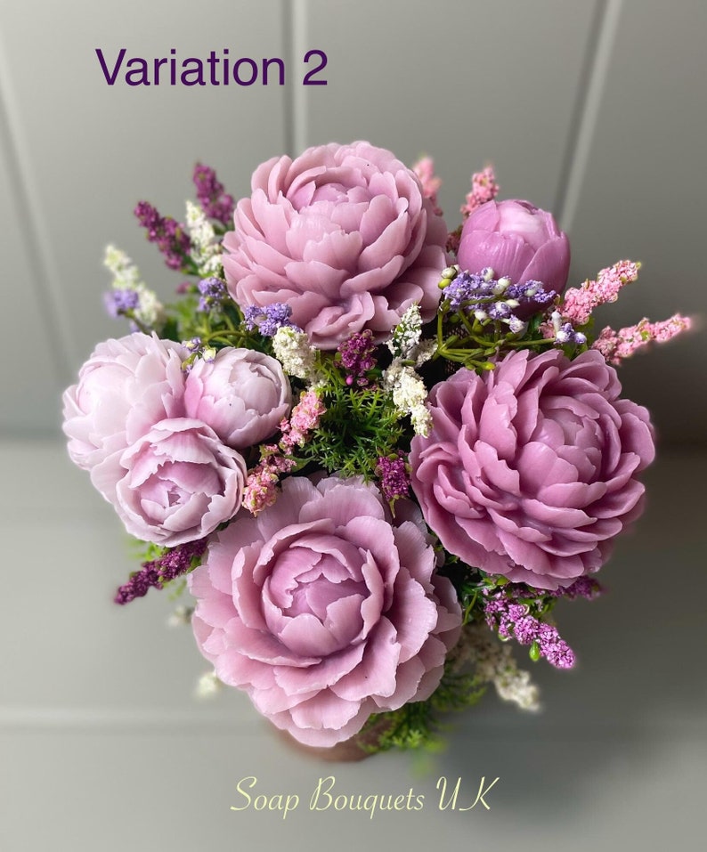 May include: A bouquet of purple soap flowers with green leaves and small pink flowers. The bouquet is arranged in a vase and the text "Soap Bouquets UK" is visible at the bottom of the image.
