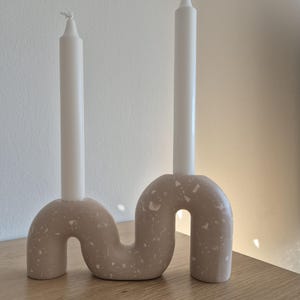 May include: A beige ceramic candle holder with a unique, abstract design. The holder has two arms that curve upwards, each holding a white candle.