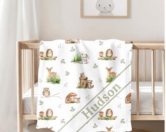 May include: White baby blanket with a woodland animal pattern, including deer, rabbits, hedgehogs, and owls. The blanket has a diagonal stripe with the name "Hudson" in green. The blanket is draped over a wooden crib.
