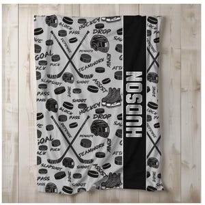 May include: A gray and black fleece blanket with a hockey theme. The blanket features hockey sticks, pucks, and other hockey-related imagery. The name "HUDSON" is printed in white on a black stripe down the center of the blanket.