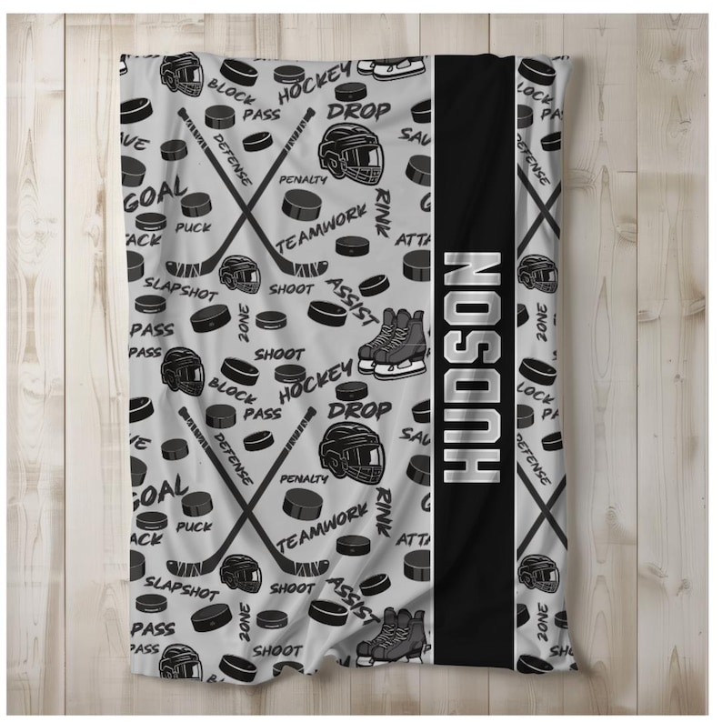 May include: A gray and black fleece blanket with a hockey theme. The blanket features hockey sticks, pucks, and other hockey-related imagery. The name "HUDSON" is printed in white on a black stripe down the center of the blanket.