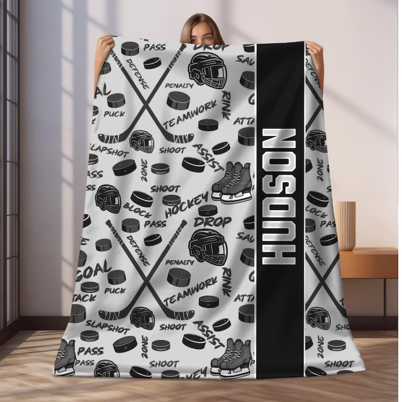May include: A gray and black fleece blanket with a hockey theme. The blanket features a repeated pattern of hockey sticks, pucks, and hockey skates. The name "HUDSON" is printed in black on a white background.