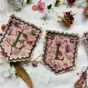 May include: A decorative banner displaying the letters "E" and "L". The pink fabric is adorned with embroidered mushrooms, branches, and crystals. The banner has green and pink scalloped edges and is strung on a white cord, creating a whimsical aesthetic.