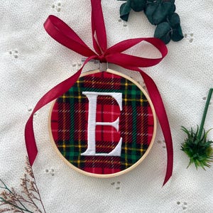 May include: A circular Christmas decoration featuring a red tartan design and a white embroidered letter "E". The design is set within a wooden hoop and has a red ribbon for hanging. Greenery is arranged around the ornament.