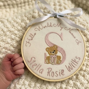 以下が含まれることがあります： 「Hello World, My Name Is Stella Rosie Wilks」の文字が刺繍されたフープアート。ピンク色の「S」の文字とテディベアのデザイン。上部には白いリボンが結ばれています。フープは木製です。