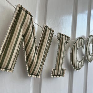 Peut inclure: Une bannière en tissu épelant "NICO". Chaque lettre est faite de tissu à rayures verticales vertes et beiges, soulignée d'une couture décorative. La bannière est suspendue à une ficelle blanche.