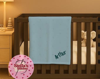 May include: A light blue baby blanket with the name "Arthur" embroidered in a darker blue font. The blanket is in a dark brown crib with a stuffed elephant toy. The blanket has a scalloped edge and a dotted pattern.