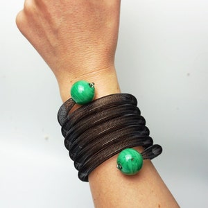 May include: A black mesh bracelet with two green malachite beads. The bracelet is wrapped around a person's wrist.