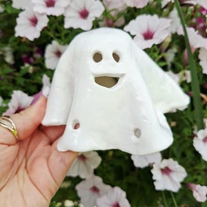 May include: A white ceramic ghost figurine with a glossy finish, featuring two circular eye cutouts and a wide, open mouth cutout. The ghost has a draped, sheet-like appearance with two additional small circular holes near the bottom edge.