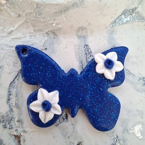 Peut inclure: Un objet décoratif en forme de papillon bleu pailleté avec deux fleurs blanches. Le papillon a un trou pour l'accrocher. Le fond est une surface marbrée blanche et grise avec des paillettes argentées.