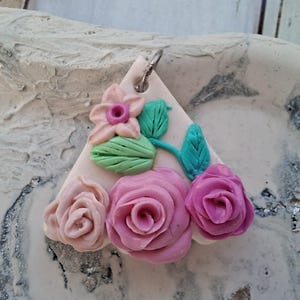 Könnte beinhalten: Ein dreieckiger Anhänger mit einer cremefarbenen Basis, die mit skulpturalen Blumenelementen verziert ist. Das Design umfasst eine kleine pfirsichfarbene Blume, zwei hellgrüne Blätter und drei Rosen in den Farben Hellrosa, Mittelrosa und Dunkelmagenta.