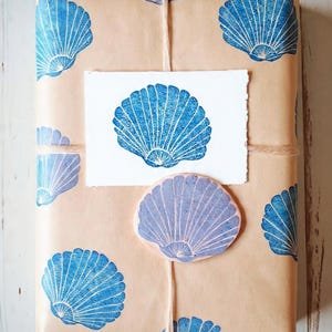 Può includere: Un regalo avvolto in carta marrone con stampe di conchiglie blu. Sono allegati un biglietto bianco e un adesivo a forma di conchiglia. Il pacco è legato con spago, creando un tema nautico.