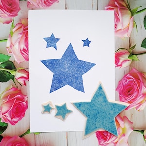 May include: A white card featuring three blue star stamps and three small star-shaped ceramic tiles. The large star on the card is flanked by two smaller stars. The ceramic stars are light blue with a textured finish. Pink roses surround the card.