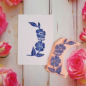May include: A blue floral design is stamped on white paper, with a matching stamp beside it. The stamp and print feature a curved arrangement of flowers and leaves. Pink roses surround the paper and stamp on a light wood background.
