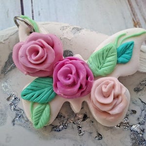 Peut inclure: Un porte-clés en forme de papillon fait à la main avec un motif floral. Les ailes sont blanches, ornées de roses roses et de feuilles vertes. Le porte-clés a un anneau argenté pour l'attache. Les fleurs sont de différentes nuances de rose.