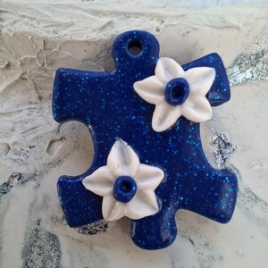 Peut inclure: Un pendentif en forme de pièce de puzzle bleu pailleté avec deux fleurs blanches. Les fleurs ont un centre bleu. Le pendentif a un trou en haut pour une chaîne. Le fond est une surface texturée blanche et argentée.
