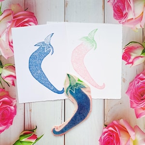 Puede incluir: Un sello de pimiento chile azul y rosa con dos impresiones en papel blanco. Las impresiones son pimientos chile azules y rosas. El sello está rodeado de rosas rosas sobre una superficie de madera blanca.