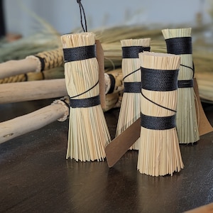 May include: A collection of small, handcrafted brooms. The brooms feature natural straw bristles bound with black twine. Each broom has a small, rectangular tag attached. The background includes larger brooms with wooden handles.