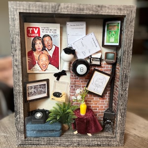 May include: A shadow box diorama with a vintage TV theme. It features a TV Guide magazine, a miniature television, an 8-ball, and other retro items. The frame is gray and the background is a brick wall.