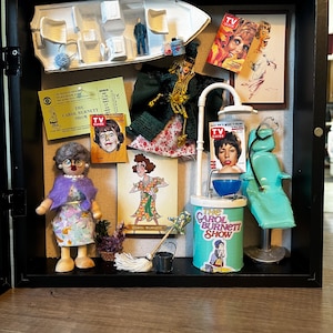 Puede incluir: Una caja de sombra negra con artículos en miniatura relacionados con The Carol Burnett Show. Incluye un barco en miniatura, portadas de revistas, una muñeca y una fregona y un cubo en miniatura.