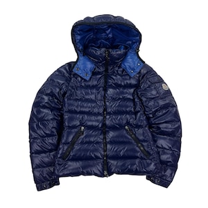 Può includere: Un piumino blu navy con cappuccio e una texture trapuntata lucida. La giacca ha una fodera blu a contrasto nel cappuccio e nel colletto, con cerniere nere e una piccola toppa con logo sulla parte superiore del braccio. La giacca è progettata per il clima freddo.