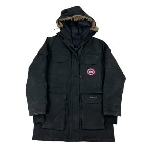 May include: A black Canada Goose parka featuring a hood with a fur trim, a front zipper closure, and multiple utility pockets. The jacket displays a circular red, white, and blue Canada Goose patch on the left chest and a small brand label on the pocket.