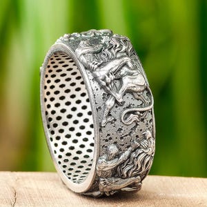May include: A silver ring featuring an intricate design of several figures, potentially mythical creatures, encircling the band. The ring has a textured surface and a perforated pattern on the inner side.