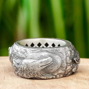 Op de afbeelding: Een zilveren ring met een gedetailleerd wolfontwerp. De wolf is afgebeeld in een slaaphouding met zijn kop rustend op zijn poten. De ring heeft een getextureerd oppervlak en een zwart diamantpatroon aan de binnenkant.