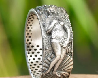 Op de afbeelding: Een zilveren ring met een gedetailleerd reliëf van een klassieke vrouwelijke figuur, mogelijk Venus, staand op een zeeschelp. De ring heeft een getextureerd oppervlak en een geperforeerde binnenband. De achtergrond is wazig, met groen gebladerte en een houten oppervlak.