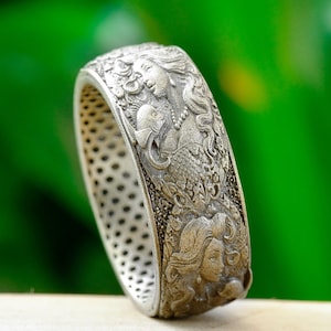 May include: A silver-toned metal ring featuring intricate relief carvings of mermaids and aquatic elements along the outer band. The inner surface of the ring displays a uniform pattern of small, circular indentations throughout the entire circumference.