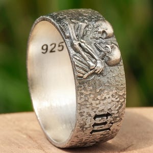 May include: A silver ring with a textured band and a detailed design of two skeletons embracing. The ring is engraved with the number "925" and the word "III".