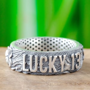 Pode incluir: Um anel de prata com as palavras "LUCKY 13" gravadas. O anel tem uma superfície texturizada e um design de trevo no lado.