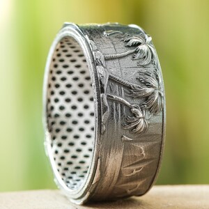 Puede incluir: Un anillo plateado con un diseño detallado de palmeras. El anillo presenta una superficie texturizada y una serie de pequeños agujeros en la banda interior. El anillo está sobre un fondo verde borroso.