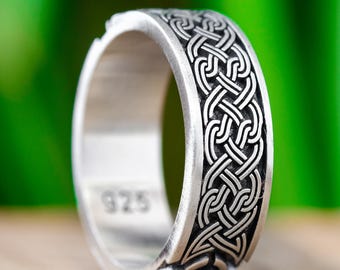 May include: A silver ring with a Celtic knot design. The ring has a detailed pattern of interwoven lines and the number "925" is visible on the inside of the band. The ring is set against a blurred green background.