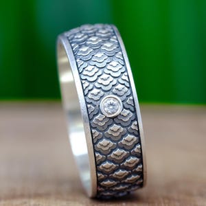 Può includere: Un anello d'argento con un motivo strutturato a scaglie attorno alla fascia. Una piccola gemma trasparente è incastonata nel design. L'anello è esposto su una superficie di legno con uno sfondo verde sfocato.