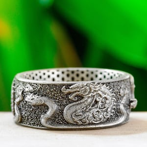 May include: A wide silver-toned metal ring featuring a textured, oxidised finish. The band is decorated with a continuous relief pattern of coiled, serpentine creatures with detailed scales and heads, wrapping around the entire circumference.