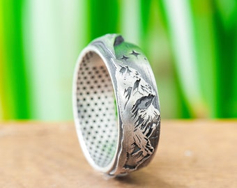 Puede incluir: Un anillo de plata con un diseño de cordillera. El anillo es texturizado y tiene un diseño detallado de montañas y estrellas.