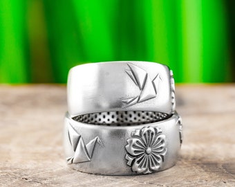 May include: Two silver rings with a textured finish. The top ring features a raised origami bird design. The bottom ring features a raised flower design.