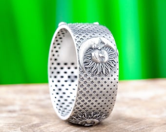 May include: A silver-toned ring with a celestial design. The ring features a sun and moon motif, surrounded by stars. The band is detailed with small, evenly spaced holes. The ring is displayed on a wooden surface against a blurred green background.