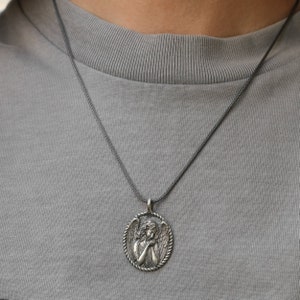 May include: A silver-toned pendant necklace featuring an oval pendant with a detailed relief of an angel. The angel is depicted with folded hands and wings, surrounded by a rope-like border. The necklace is worn on a gray shirt.