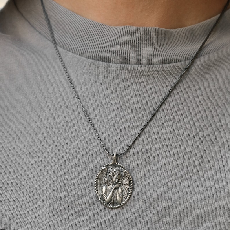 May include: A silver-toned pendant necklace featuring an oval pendant with a detailed relief of an angel. The angel is depicted with folded hands and wings, surrounded by a rope-like border. The necklace is worn on a gray shirt.