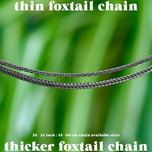 May include: Two silver foxtail chains are displayed against a blurred green background. The text "thin foxtail chain" and "thicker foxtail chain" are at the top and bottom. Available sizes are 20-24 inches.