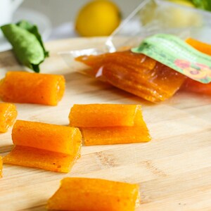 May include: A wooden cutting board with several orange fruit leather rolls. The rolls are arranged in a row and are slightly overlapping. There is a small green leaf in the background.