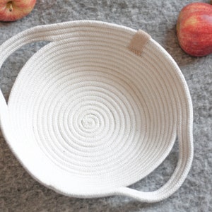 May include: A white, round, rope basket with two handles. The basket is made of thick, white rope and has a woven pattern.