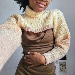 May include: Crocheted bolero sweater in cream, beige, and light brown. The sweater features a turtleneck and long sleeves with a ribbed cuff. The color gradient transitions from cream to light brown. The bolero is a cropped style.