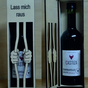 Puede incluir: Una caja de madera para botella de vino con un diseño de celda de prisión. La caja tiene un recorte de manos sosteniendo los barrotes con el texto "Lass mich raus" en la parte delantera. La caja está abierta y una botella de vino Castiza Tempranillo está dentro.